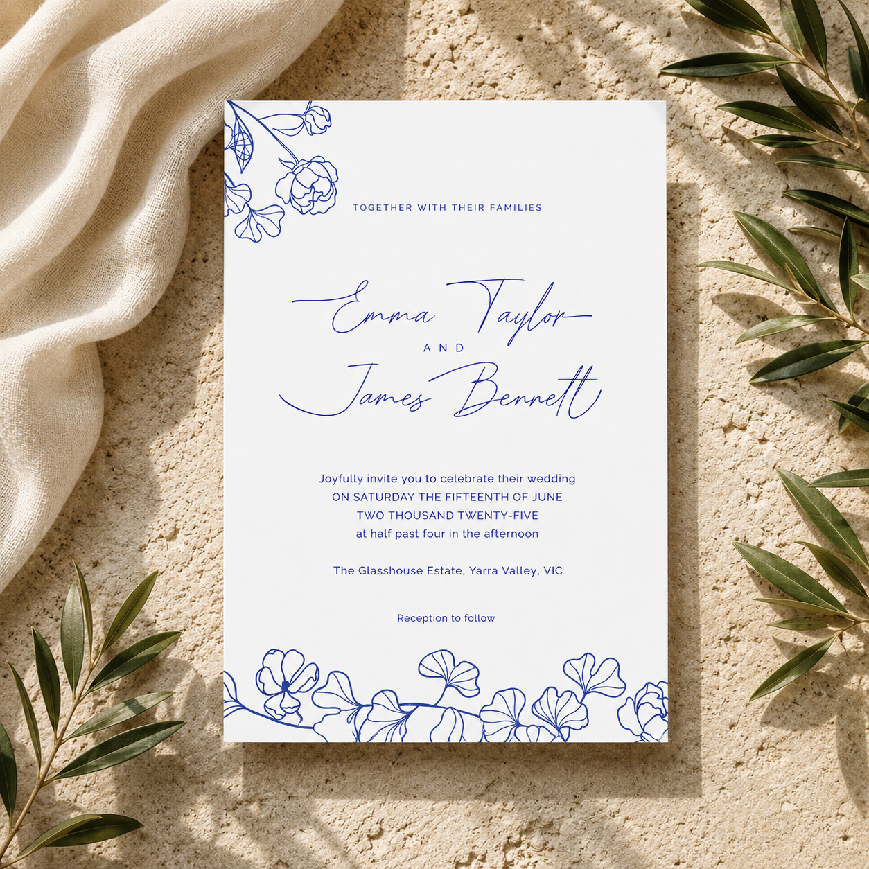 A wedding invitation card with blue floral line art at the top and bottom and elegant script names, photographed flat on a textured beige surface with draped cream fabric and green leaves around it.