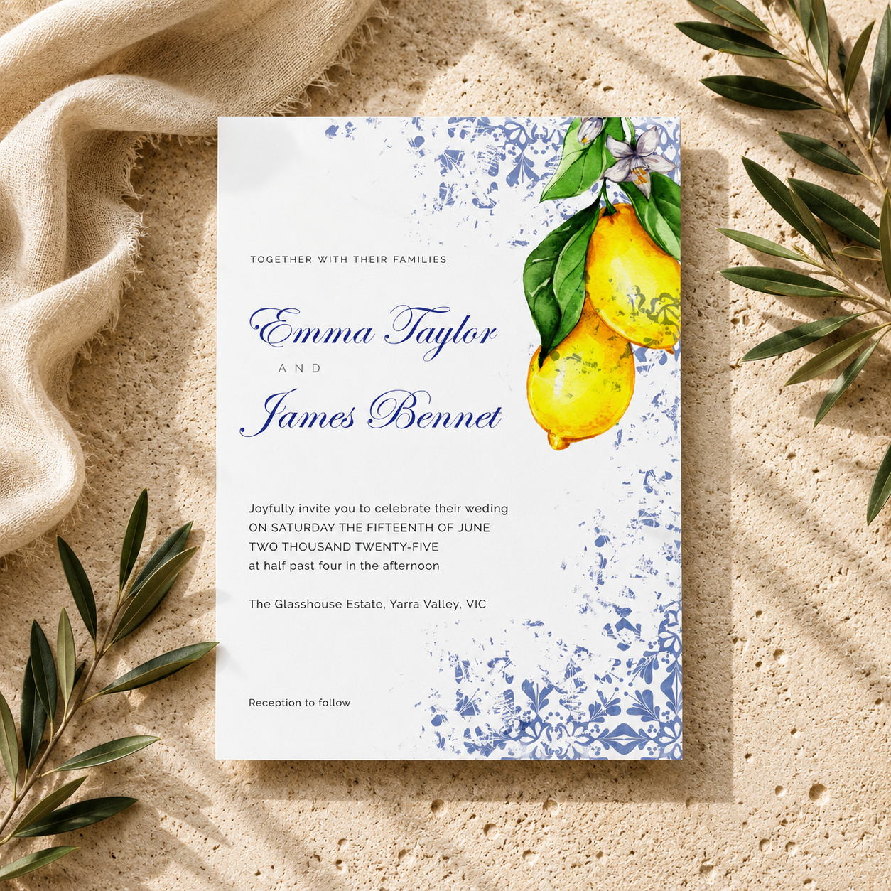 A square wedding invitation card with a white background, blue floral border, and yellow lemons with green leaves, styled on a beige textured surface with draped fabric and olive branches.