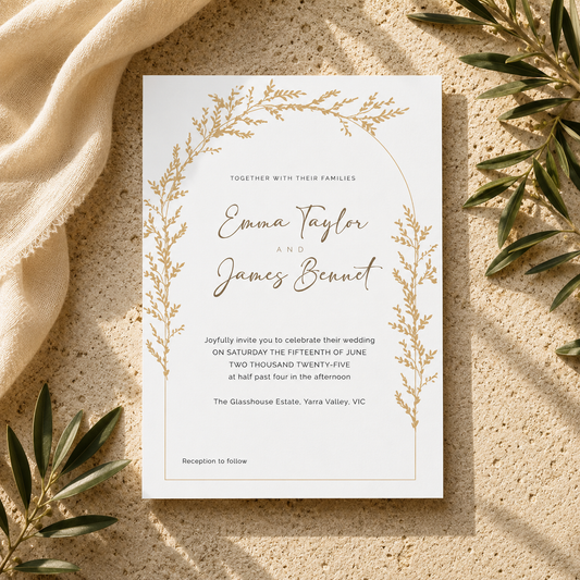A vertical wedding invitation card with a delicate gold botanical wreath border and elegant script text, styled flat-lay on a textured beige stone surface with cream fabric and olive branches.