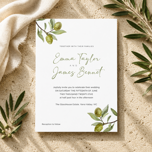 A wedding invitation card with olive branch illustrations, styled flat-lay on a sandy surface with a beige fabric and green leaves.