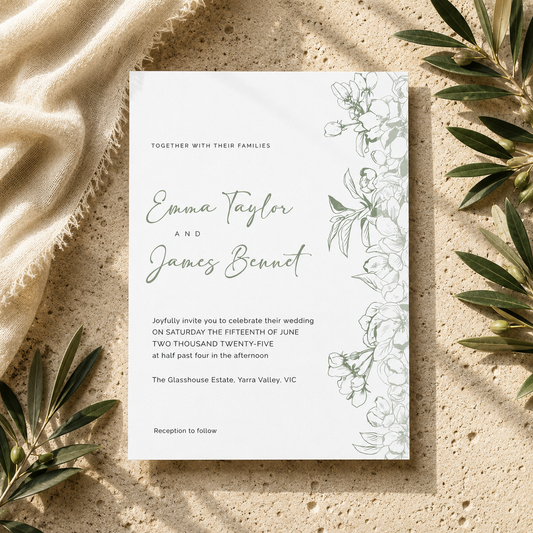 A wedding invitation card with elegant green floral illustrations on the right side, laid flat on a textured beige stone surface with draped cream fabric and olive leaves around it.