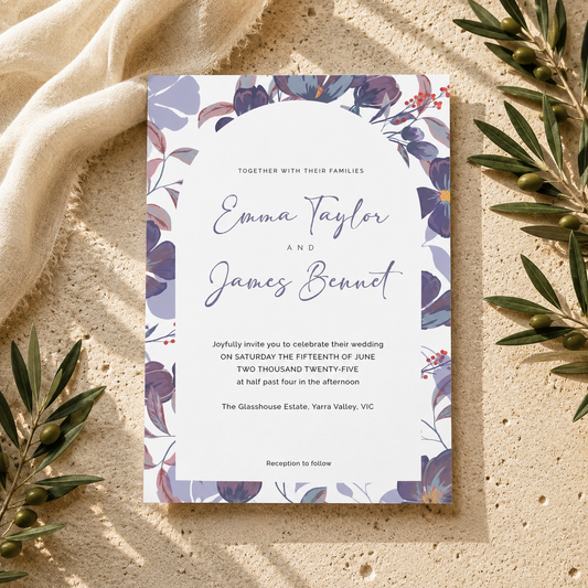 A floral wedding invitation card with purple and blue botanical borders, styled flat-lay on a textured beige surface with olive branches and draped fabric.