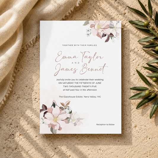 A rectangular wedding invitation card with elegant script and floral watercolor accents, styled flat-lay on a textured beige surface with draped fabric and green leaves.