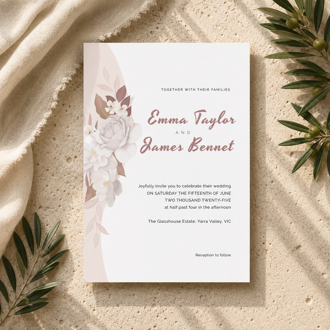 A vertical wedding invitation card with a soft blush floral design on a white background, styled flat-lay on a textured beige surface with draped fabric and green leaves.