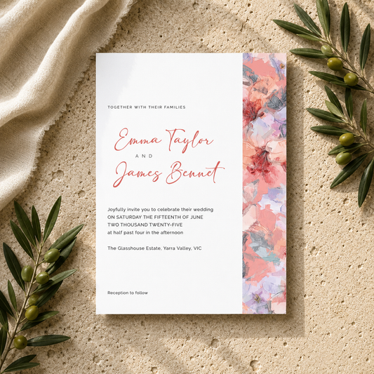 A wedding invitation card with elegant coral script text and a vertical pink, peach, and blue floral watercolor border on the right, styled flat-lay on a textured beige stone surface with draped cream fabric and olive branches.
