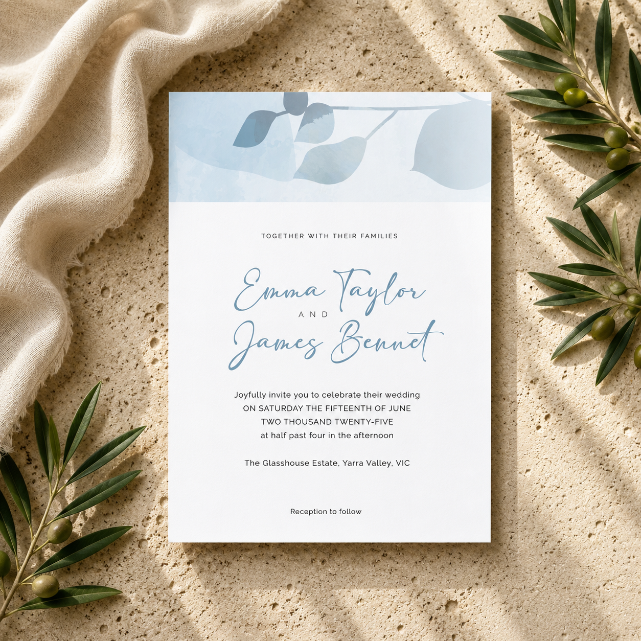 A square wedding invitation card with elegant blue script and serif text, styled flat-lay on a textured beige stone surface with draped cream fabric and olive branches.