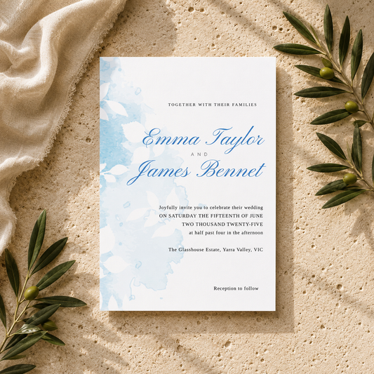 A wedding invitation card with light blue watercolor floral details and elegant blue script text, styled flat-lay on a textured beige stone surface with draped cream fabric and olive branches.