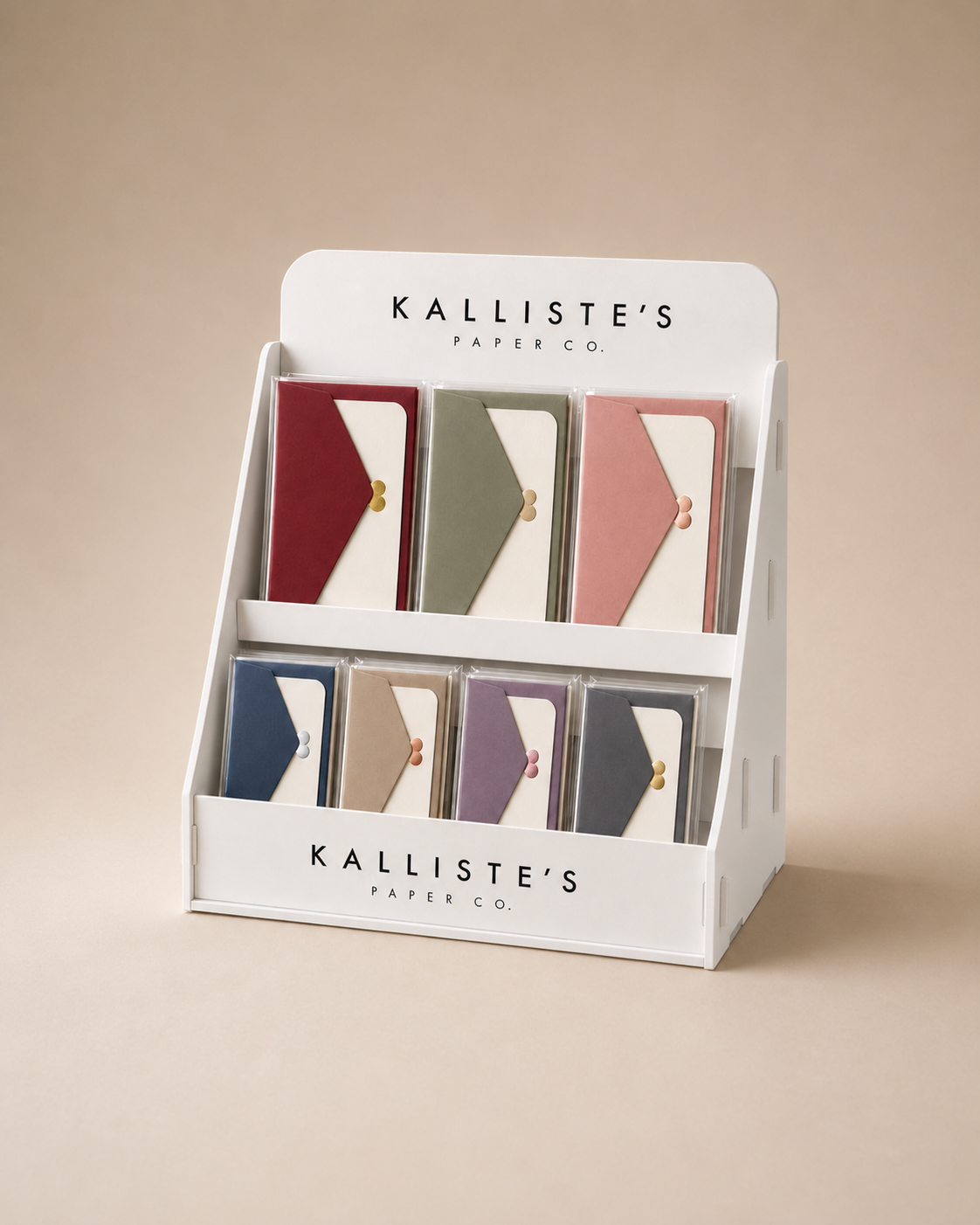 A white Kalliste's Paper Co. display stand holding multiple small paper goods packages in assorted colors, photographed against a warm beige studio background.
