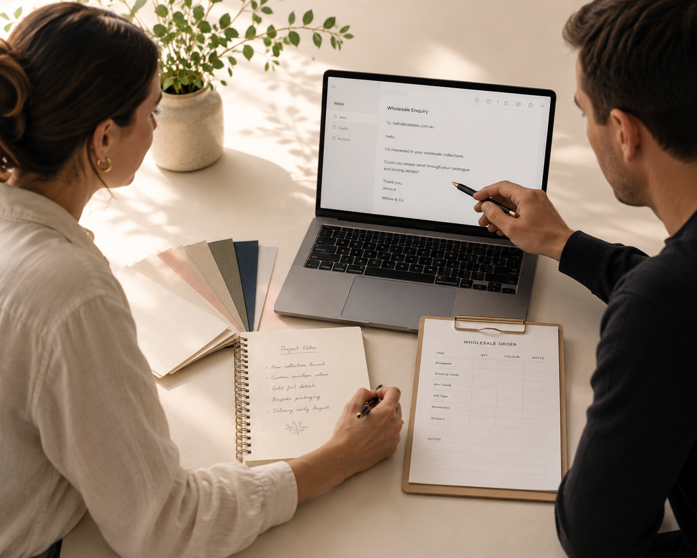 A man and a woman sit at a table looking at a laptop screen, with the woman writing in a notebook and the man pointing at the display. Papers and a small potted plant are on the table in a bright indoor setting.