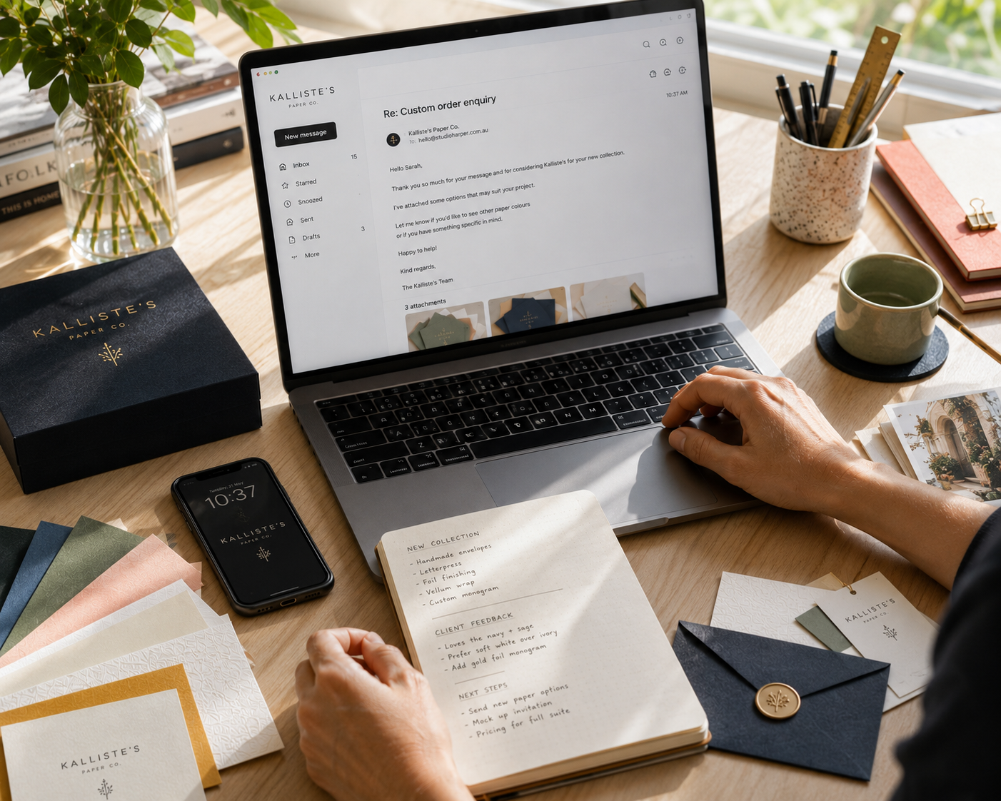 A styled desk scene with a person's hands using a laptop on a wooden table. Visible branded items include a black Kallister's Leather gift box, a black smartphone, stationery, cards, and an open notebook.