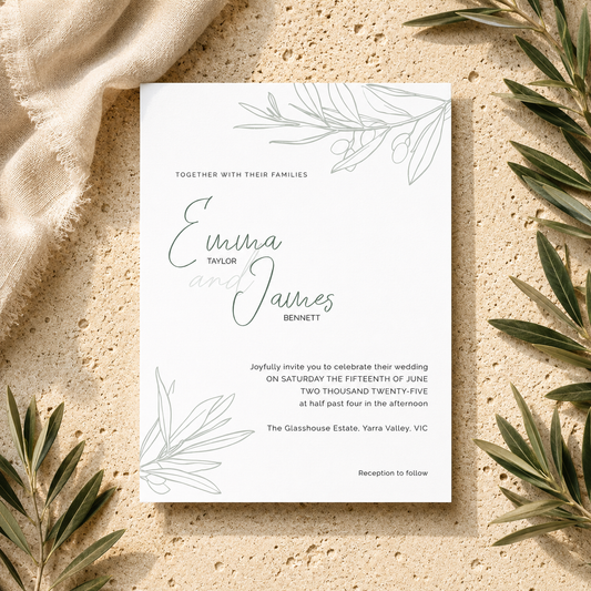 A square white wedding invitation card with delicate gray botanical line art in the corners, styled in a flat lay on a textured beige stone surface with a cream linen cloth and olive branches around the edges.