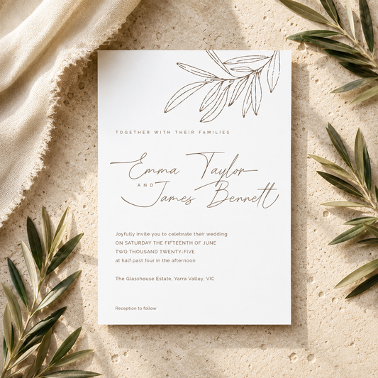 A square wedding invitation card with elegant black script and line art, styled in a flat lay on a textured beige surface with draped cream fabric and olive branches around it.