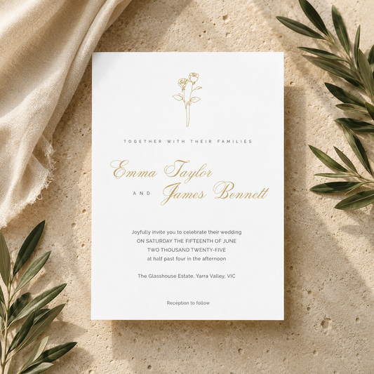 A square white wedding invitation card with elegant gold script and floral line art, styled flat-lay on a textured beige surface with draped cream fabric and olive green leaves around it.