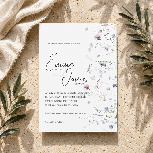 A vertical wedding invitation card with elegant black script and floral watercolor illustrations in soft purple, blue, and green, styled flat-lay on a textured beige stone surface with linen and olive branches.