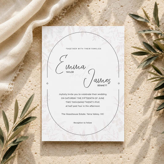 A square wedding invitation card with elegant black script text and a faint damask pattern, styled in a flat-lay on a textured beige stone surface with draped cream fabric and olive branches.