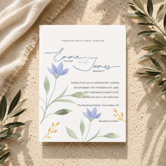 A square wedding invitation card with elegant script names and floral botanical illustrations, styled flat-lay on a textured beige surface with olive leaves and draped fabric.