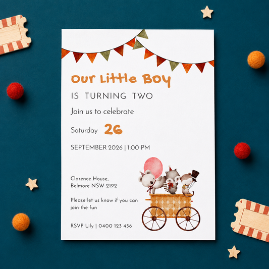 A baby birthday invitation card styled in a flat-lay on a dark teal background with circus-themed props like tickets, pom-poms, and wooden stars. The white card features orange and black text reading 'Our Little Boy IS TURNING TWO' and includes a cartoon illustration of three clown characters in a carriage.