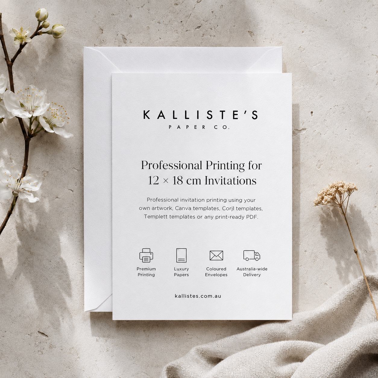 A white invitation card and matching envelope from Kalliste's Paper Co. are displayed in a styled flat-lay on a textured light stone surface with dried white flowers and a beige fabric accent.