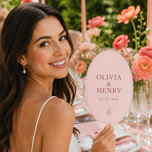 A smiling woman at an outdoor wedding table holds a personalized oval pink sign that reads 'OLIVIA & HENRY 14 - 11 - 2026' with a small floral motif. The background shows a decorated garden table with pink flowers, glassware, and soft warm lighting.