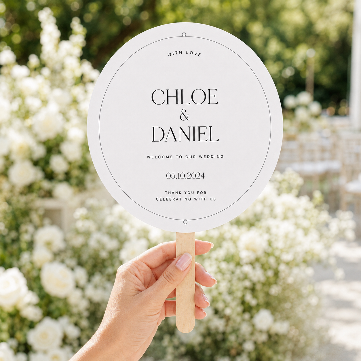 A hand holds a round white wedding fan sign with a wooden handle in front of a blurred outdoor garden background with white flowers. The sign reads 'WITH LOVE CHLOE & DANIEL WELCOME TO OUR WEDDING 05.