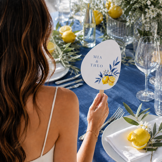 A woman at a decorated blue table setting holds a personalized oval sign that reads 'MIA & THEO' with blue floral and yellow lemon illustrations.