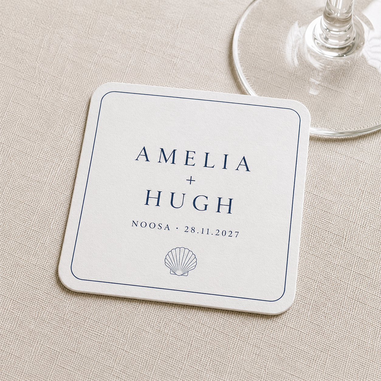 A square white personalized wedding coaster with navy blue text reading 'AMELIA + HUGH', 'NOOSA • 28.11.