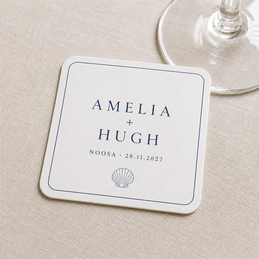 A square white personalized wedding coaster with navy blue text reading 'AMELIA + HUGH', 'NOOSA • 28.11.