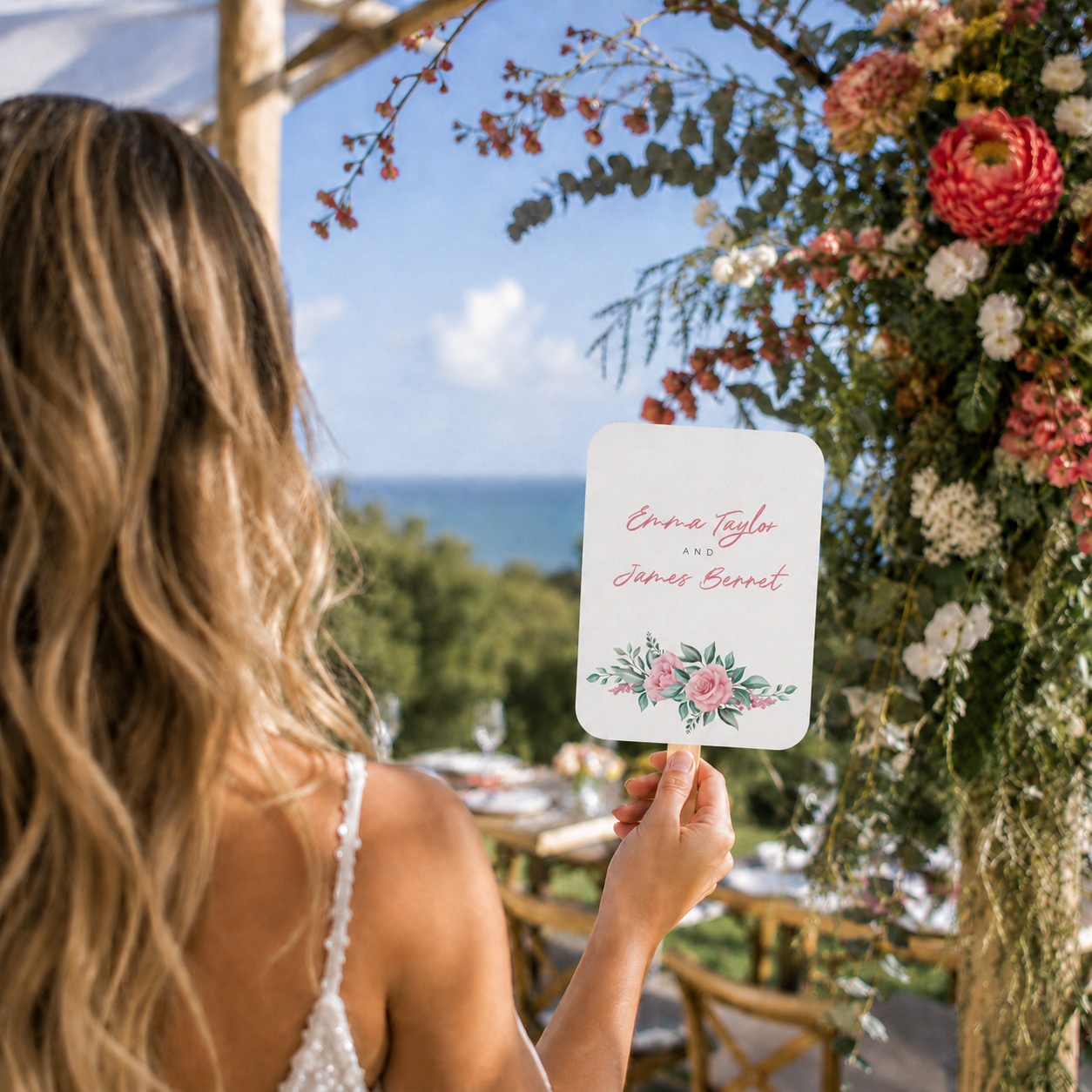 A woman at a wedding holds a custom printed sign featuring pink script text and a floral design at the bottom, set against an outdoor backdrop with a flower arch and ocean view.