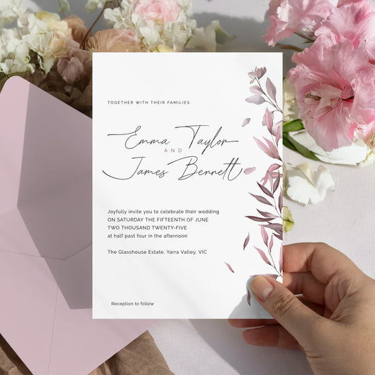 A hand holds a white wedding invitation card with elegant black script and muted pink floral illustrations on the right side. The card is displayed over a pink envelope, with soft pink and white flowers arranged in the background.