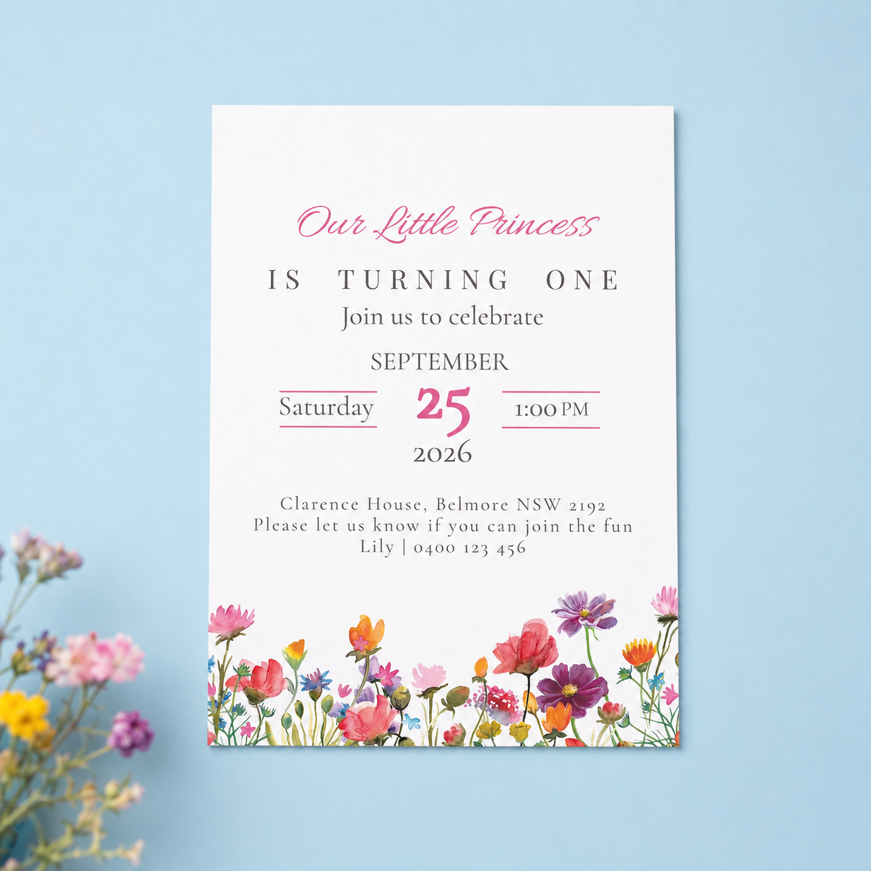 A vertical baby first birthday invitation card with a white background, pink and black text, and a colorful floral border at the bottom, displayed on a light blue background with decorative flowers in the corner.