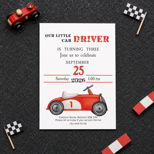 A baby racing birthday invitation card styled in a flat-lay on a dark asphalt background, surrounded by toy race elements including a red toy car, checkered flags, and red-and-white striped poles.