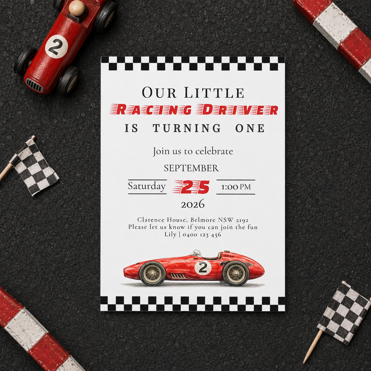 A racing-themed first birthday invitation card is displayed on a dark textured surface with toy cars, checkered flags, and striped blocks around it. The card features a black-and-white checkered border, red racing text, and an illustration of a red race car with the number 2.