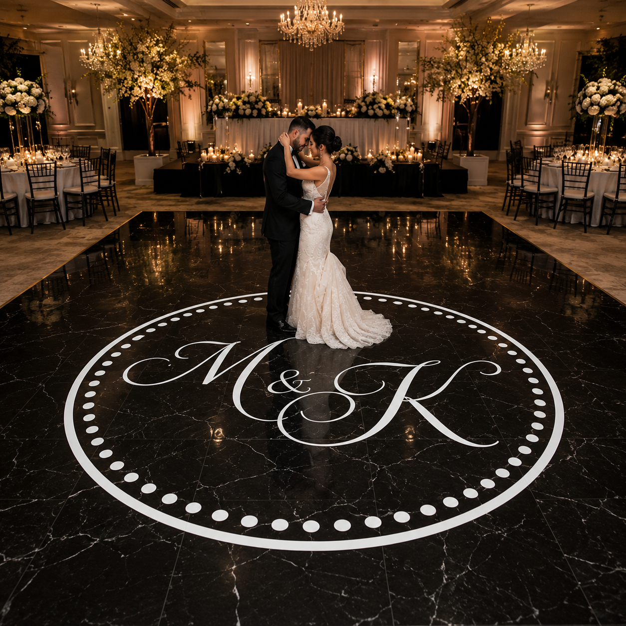 A bride in a white wedding gown and a groom in a black suit kiss on a polished black marble dance floor featuring a large white circular monogram design. The background shows a grand, elegantly decorated wedding reception hall with chandeliers, floral arrangements, and dining tables.