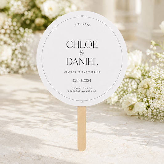 A round white wedding welcome sign on a wooden stick, printed with the names 'Chloe & Daniel' and wedding details, set against a soft-focus background of white flowers.