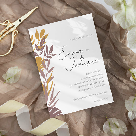 A wedding invitation card with botanical leaf illustrations in gold, mauve, and brown on a white background, styled on crumpled taupe paper with gold scissors, a cream ribbon, and white flowers.
