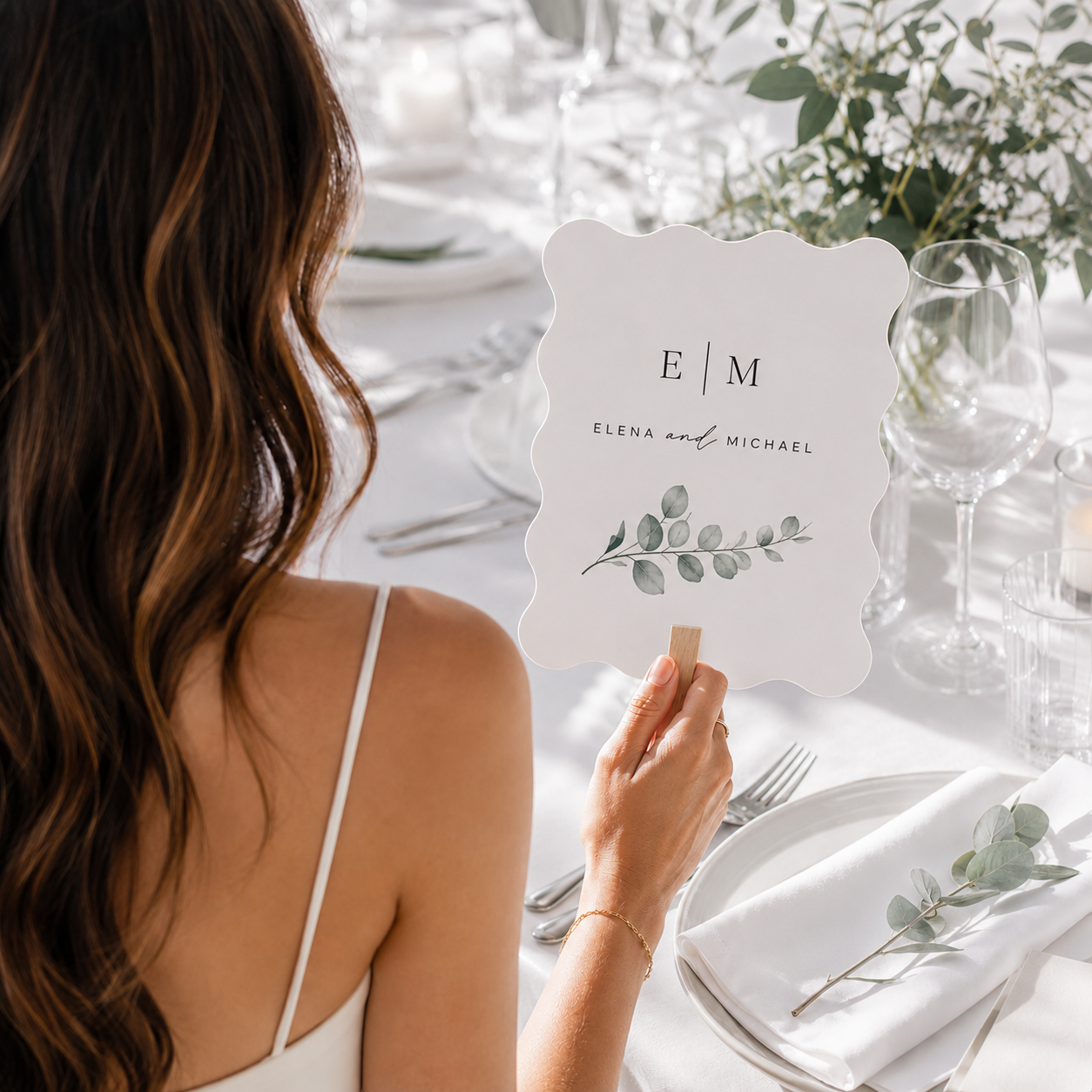 A woman at a wedding table holds a white scalloped place card with the names 'Elena and Michael' and a green eucalyptus illustration. The table is elegantly set with white linens, glassware, and floral arrangements in soft natural light.
