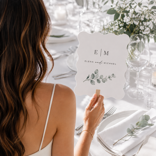 A woman at a wedding table holds a white scalloped place card with the names 'Elena and Michael' and a green eucalyptus illustration. The table is elegantly set with white linens, glassware, and floral arrangements in soft natural light.