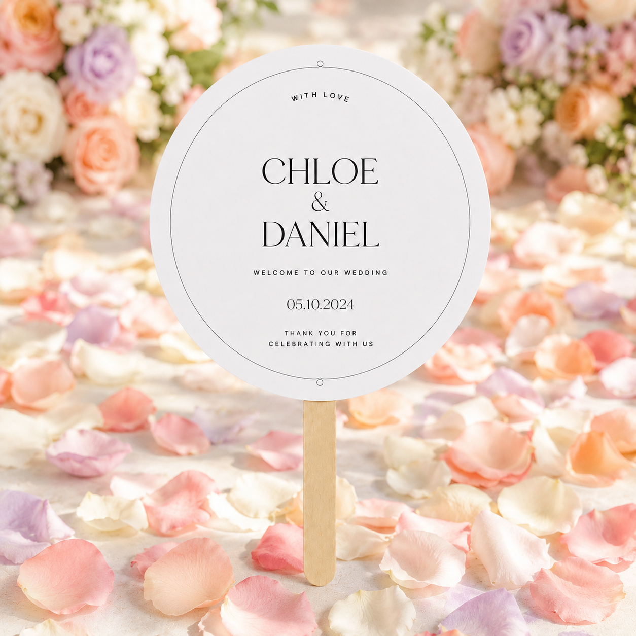 A round white wedding sign on a wooden stick, printed with the names 'CHLOE & DANIEL' and wedding details, set against a background of scattered pink and peach flower petals with blurred floral arrangements.
